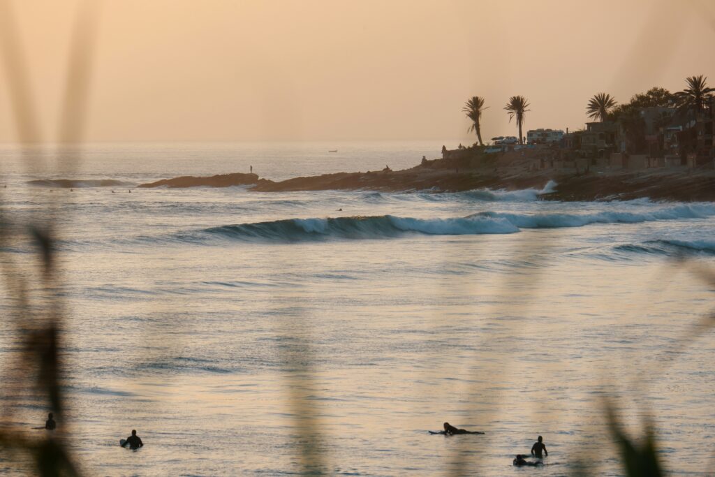 Rolling right-hand wave at Taghazout surf Beach, Morocco surf, with people learning to surf and onlookers on the shore against yellow skies.