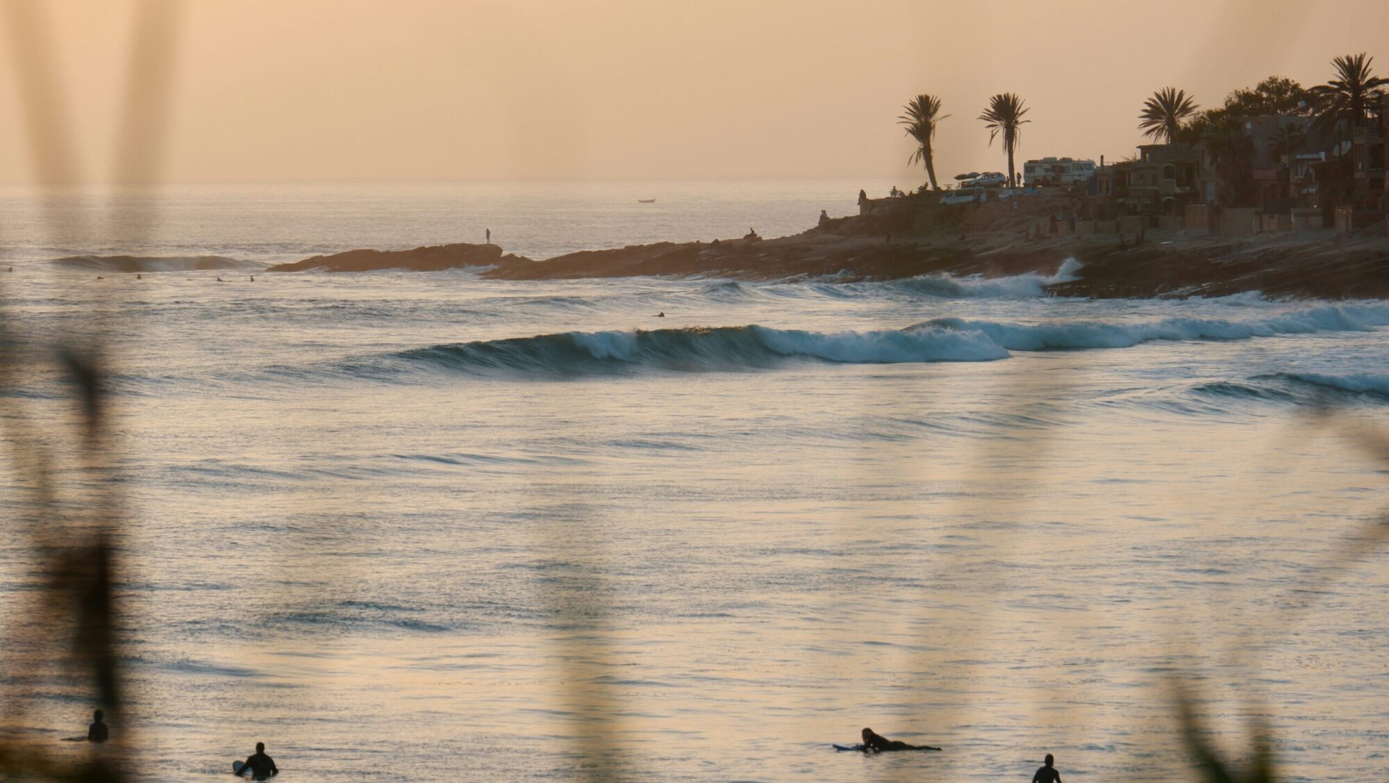 Rolling right-hand wave at Taghazout surf Beach, Morocco surf, with people learning to surf and onlookers on the shore against yellow skies.