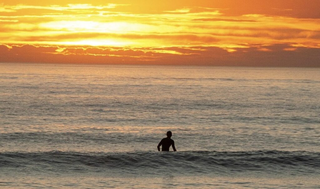 A person sitting on their surfboard in a calm ocean, with a vibrant yellow sunset illuminating the sky in the background in Mancora surf