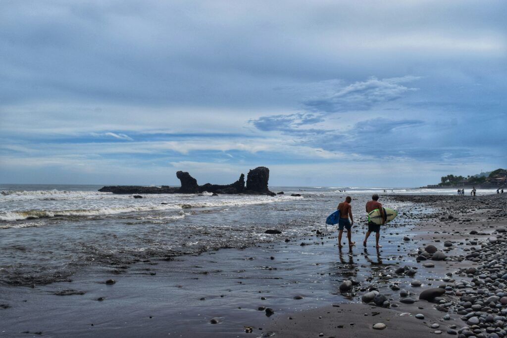 Two surfers walking down the beach at El Tunco surf, El Salvador, carrying their surfboards and navigating a rocky shoreline