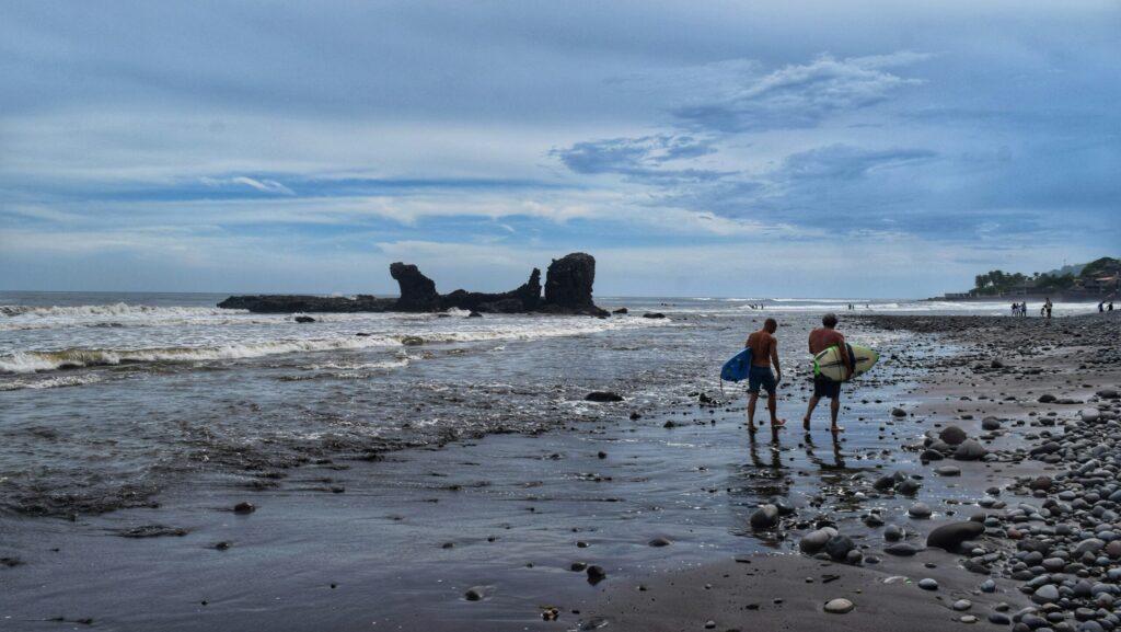 Two surfers walking down the beach at El Tunco surf, El Salvador, carrying their surfboards and navigating a rocky shoreline