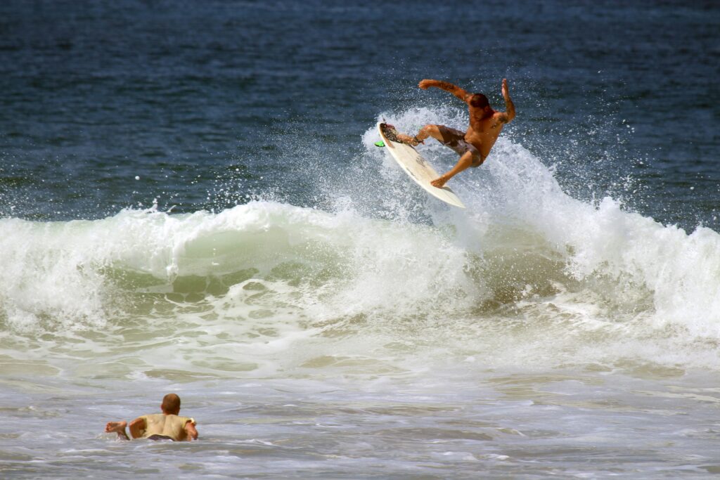 A surfer executing an aerial maneuver in Canoa Surf above the wave, with water spraying around them, while another surfer watches from nearby