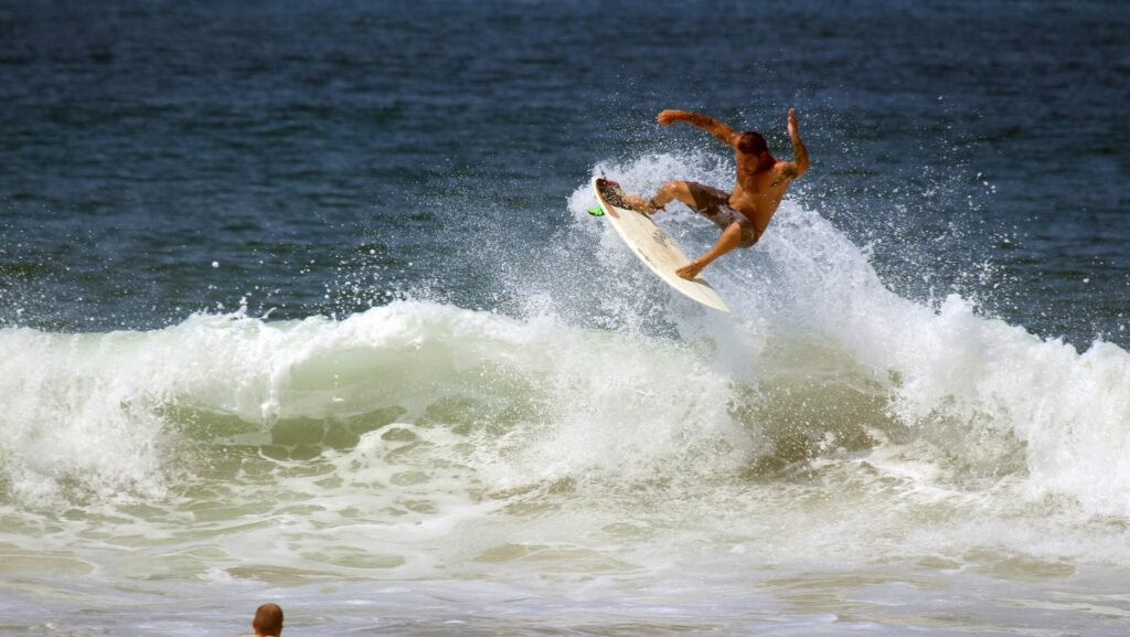 A surfer executing an aerial maneuver in Canoa Surf above the wave, with water spraying around them, while another surfer watches from nearby