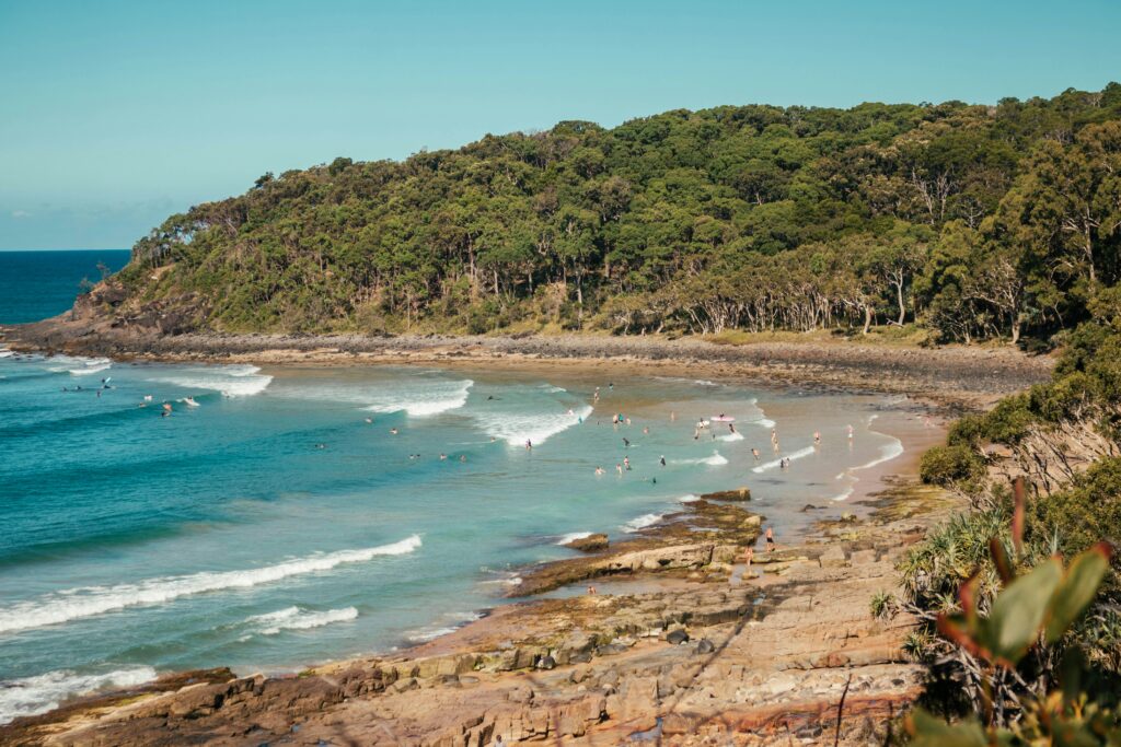 People swimming and surfing at Tea Tree Bay Surf, Noosa, with the lush backdrop of the national park