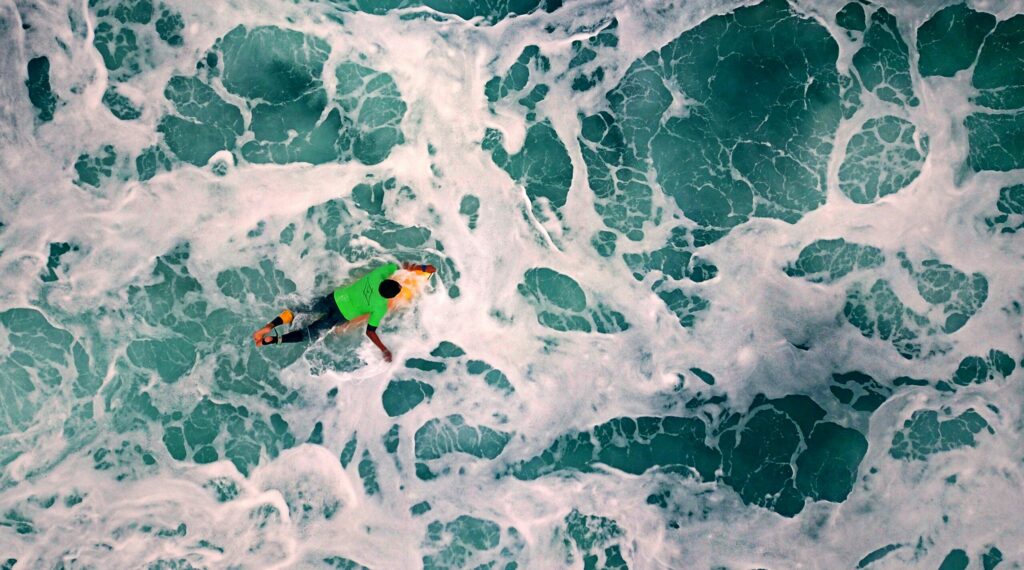 A bird's-eye view of a surfer wearing a green vest, zoomed in on the backwash of water as they navigate through the waves