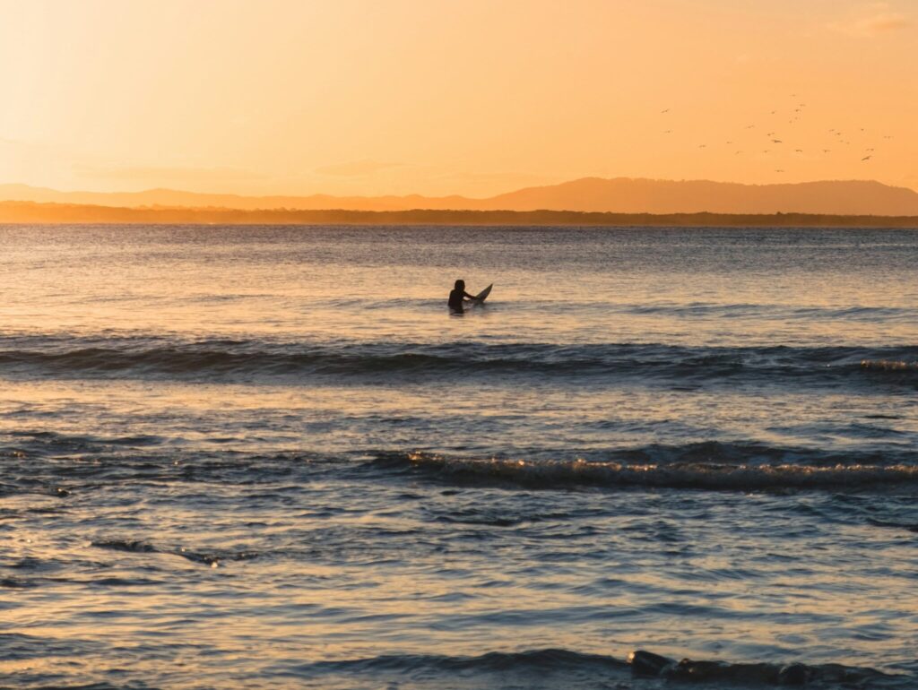 Surfer waiting for a wave at First Point, Noosa Heads, with a stunning sunset and vibrant yellow skies