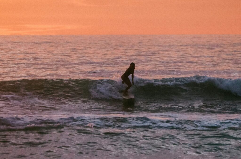 A girl surfing at Canoa surf, a small wave under a vibrant yellow and orange sky, capturing the beauty of the sunset.