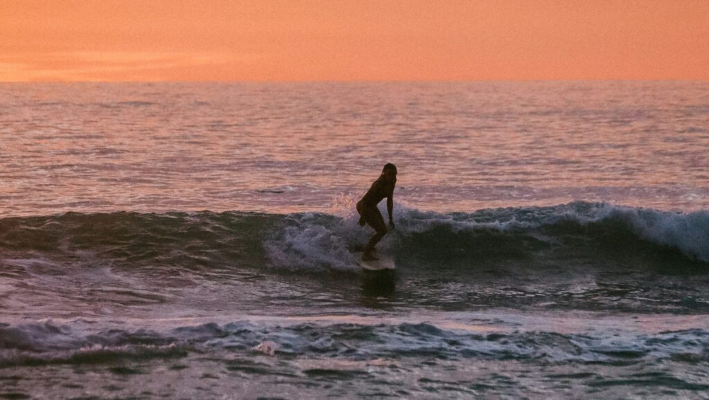 A girl surfing at Canoa surf, a small wave under a vibrant yellow and orange sky, capturing the beauty of the sunset.