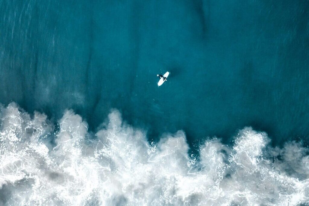 Bird's eye drone view of a single surfer in deep blue water, surrounded by waves under clear skies