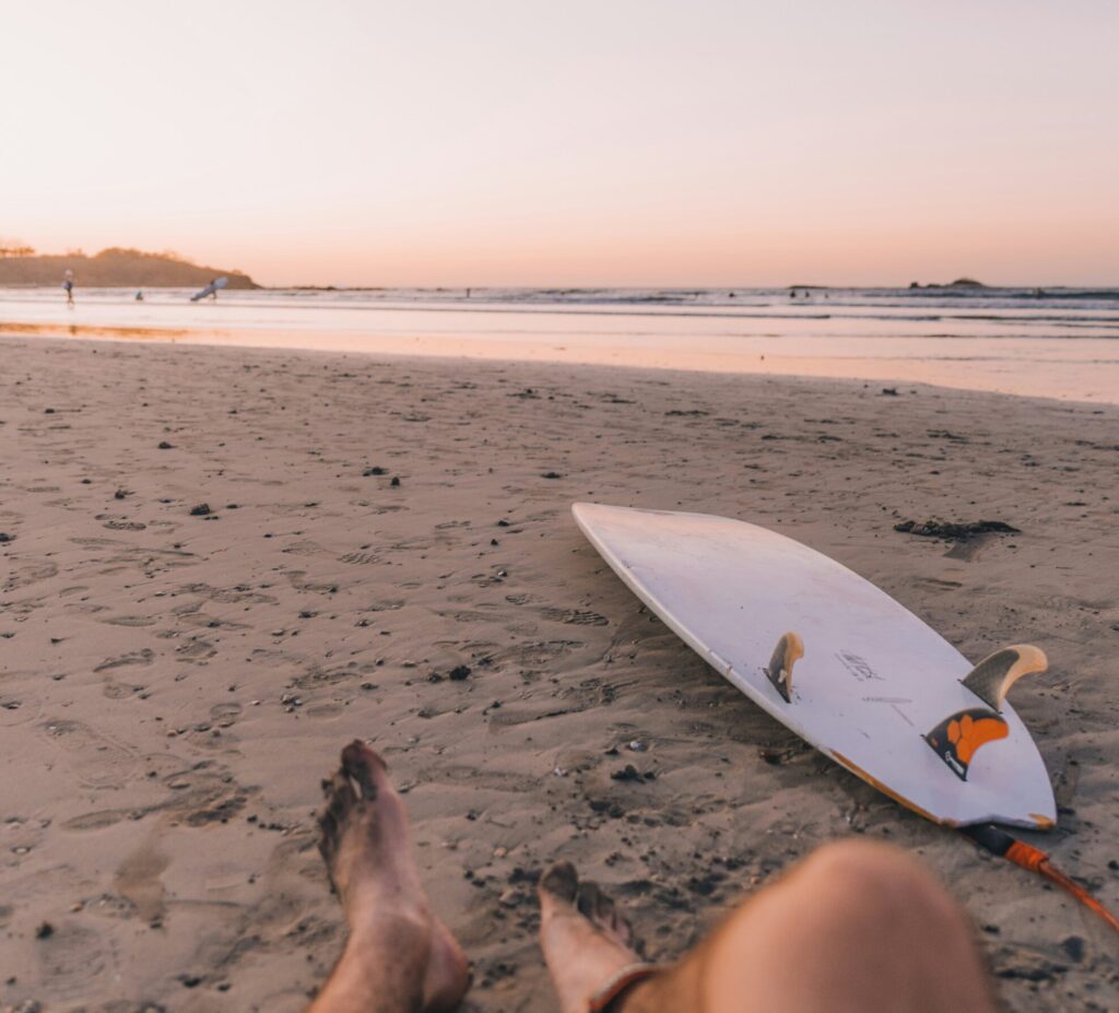 A man sitting on the sandy beach in Jaco, Costa Rica, with his surfboard beside him, gazing out at the ocean waves under a clear sky