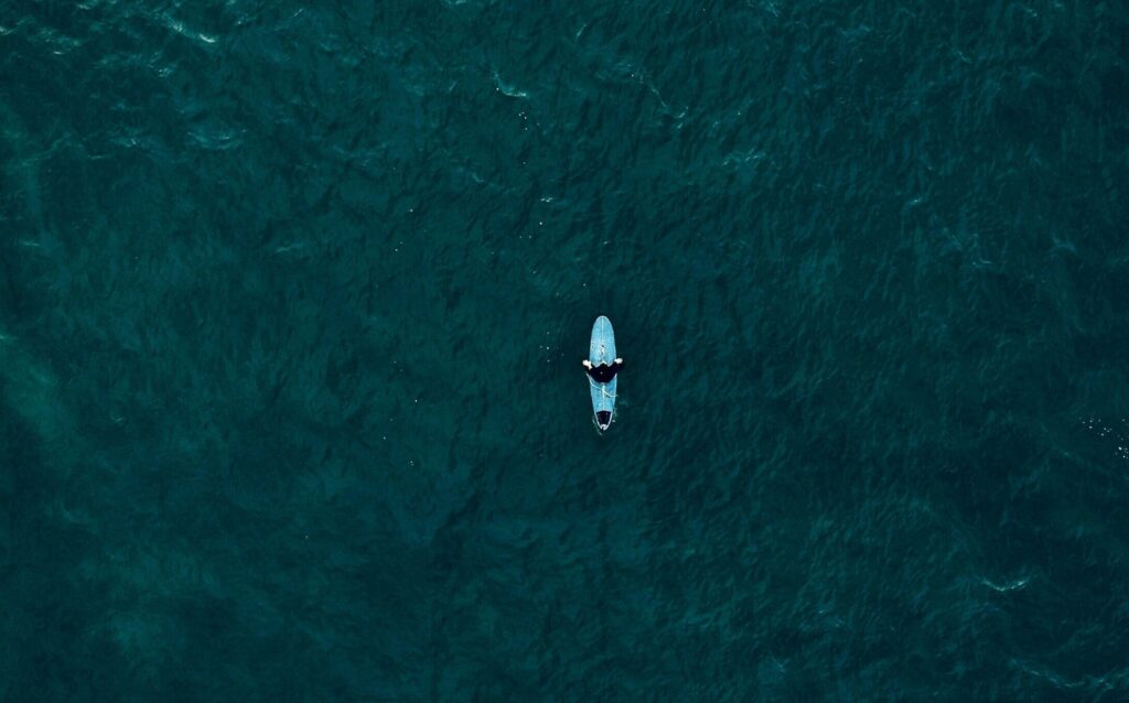 A bird's-eye view of a single surfer in deep blue, calm water, surrounded by tranquility as they float on their board