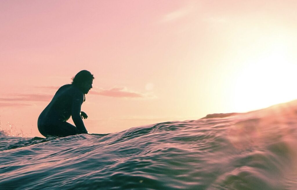 Surfer rides a wave at sunset, seen from behind in the water, with vibrant orange and yellow skies