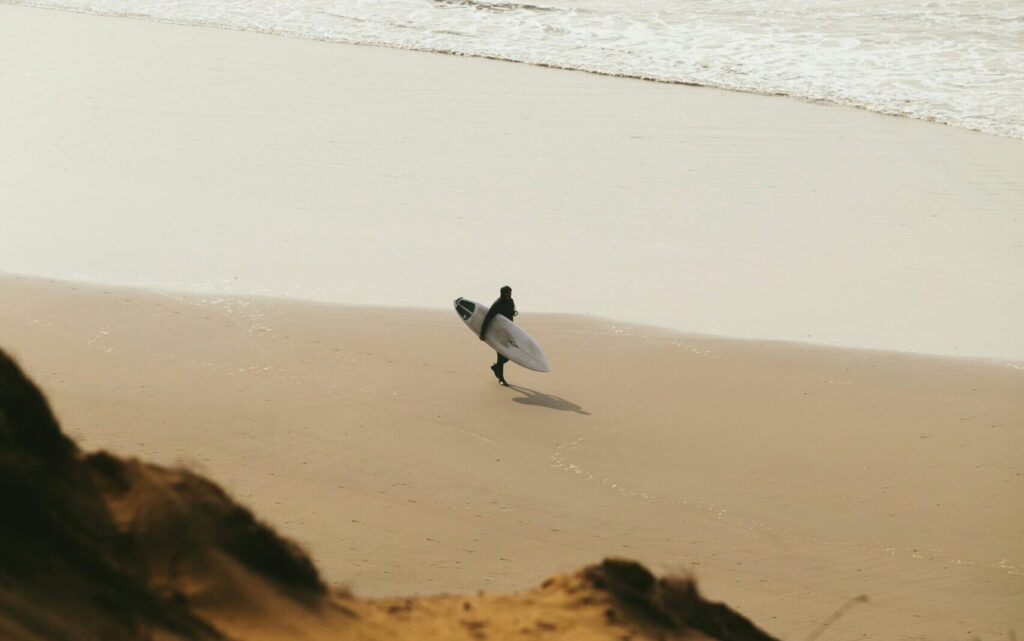 A surfer walking along the beach, captured from atop a sand dune, providing a unique perspective as they stroll along the shoreline