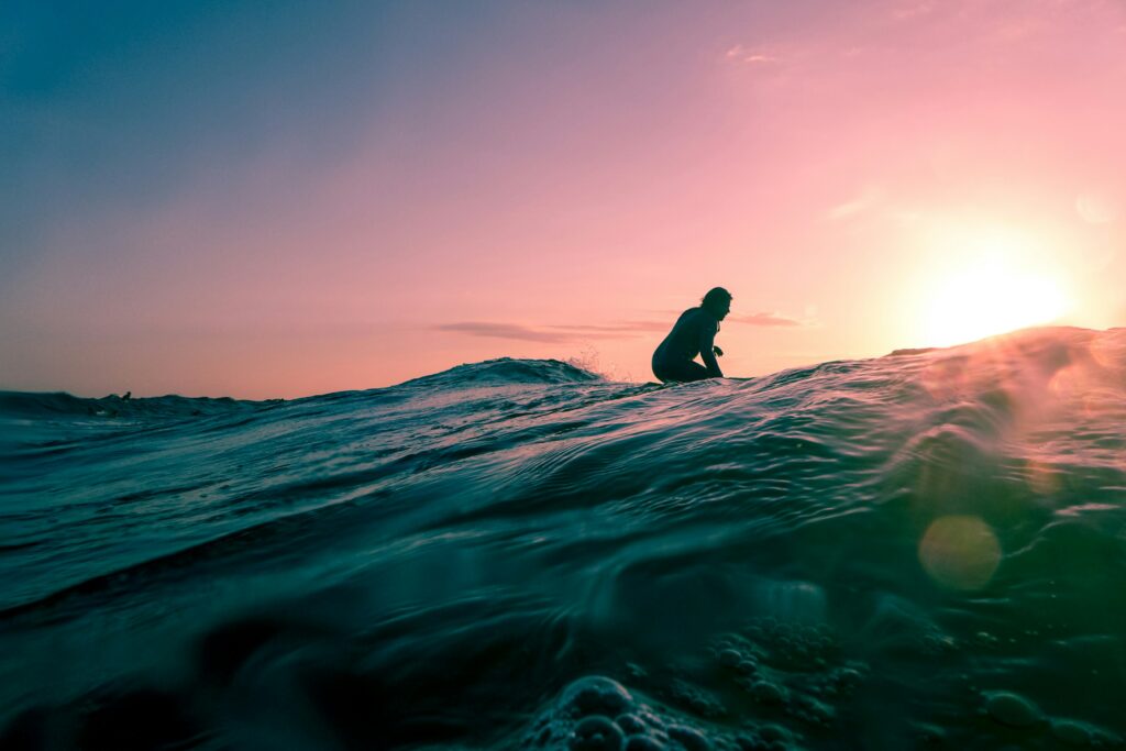 A view of a surfer from behind the wave, silhouetted against a sunset of vibrant orange, blue, and yellow hues in Canoa surf