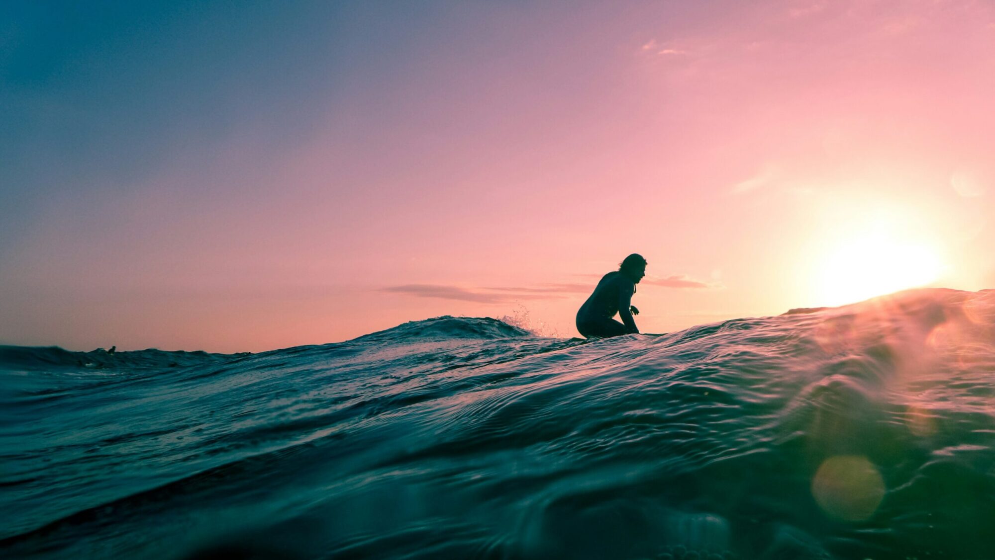 A view of a surfer from behind the wave, silhouetted against a sunset of vibrant orange, blue, and yellow hues in Canoa surf