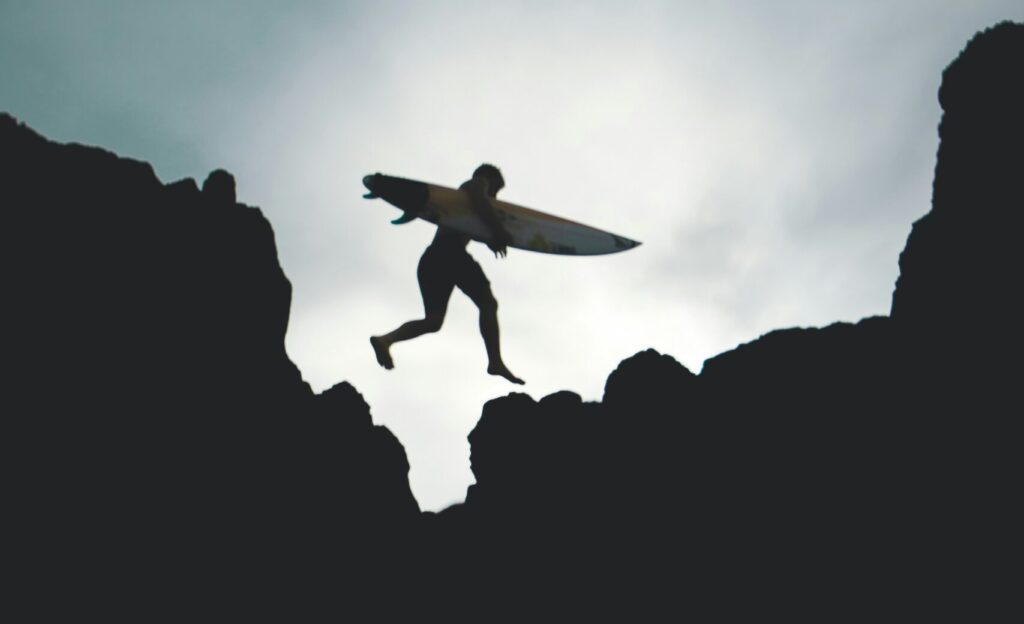 A man in black and white leaping over rocks while carrying a surfboard, captured mid-air against a dramatic coastal backdrop