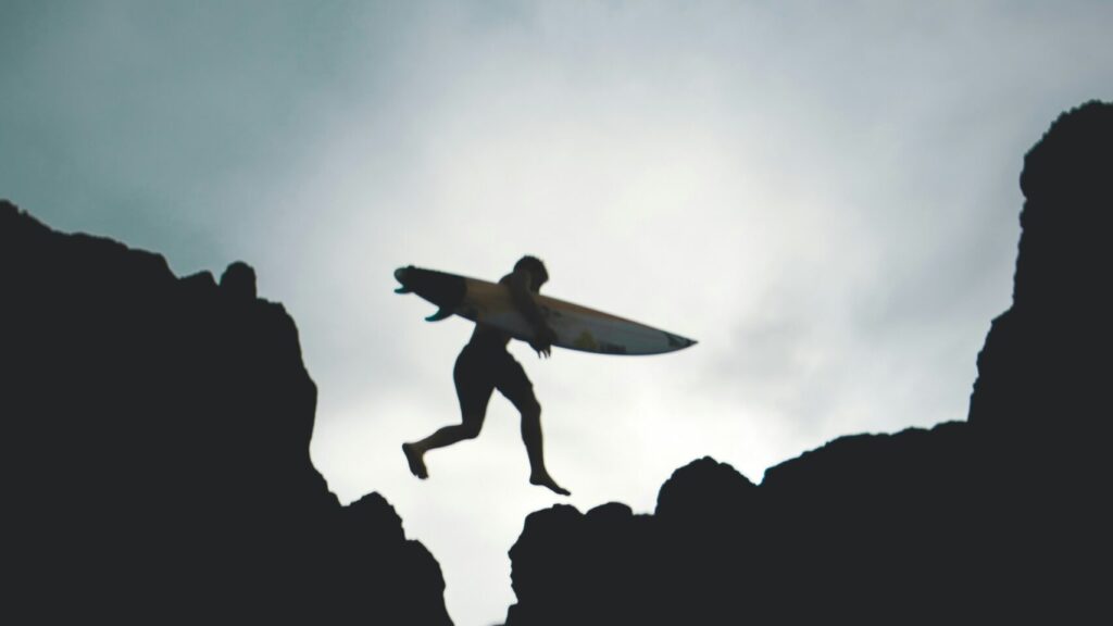 A man in black and white leaping over rocks while carrying a surfboard, captured mid-air against a dramatic coastal backdrop