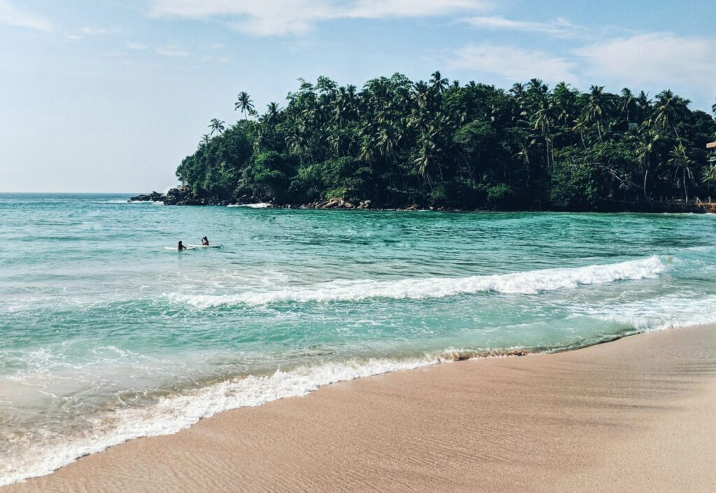 People learning to surf at Hiriketiya Beach, surrounded by clear blue water and lush tropical trees in the background