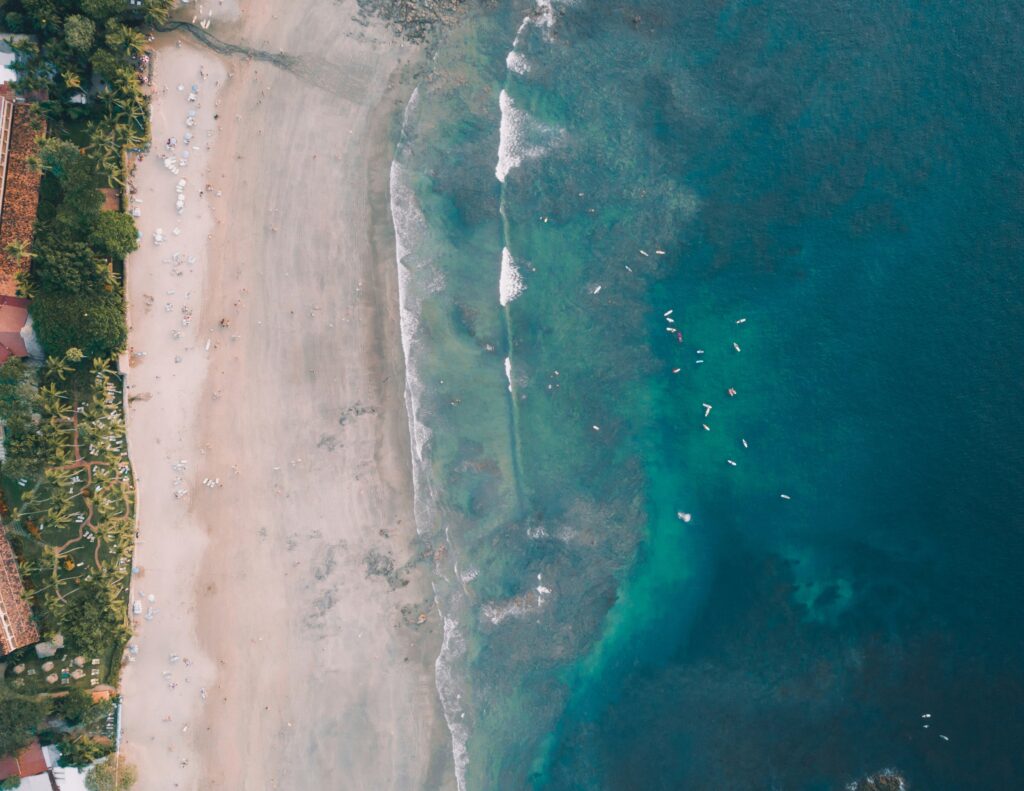 A bird's-eye view of Tamarindo Beach, Costa Rica Surf, showing surfers riding waves, a sandy beach lined with palm trees, and vibrant blue ocean waters