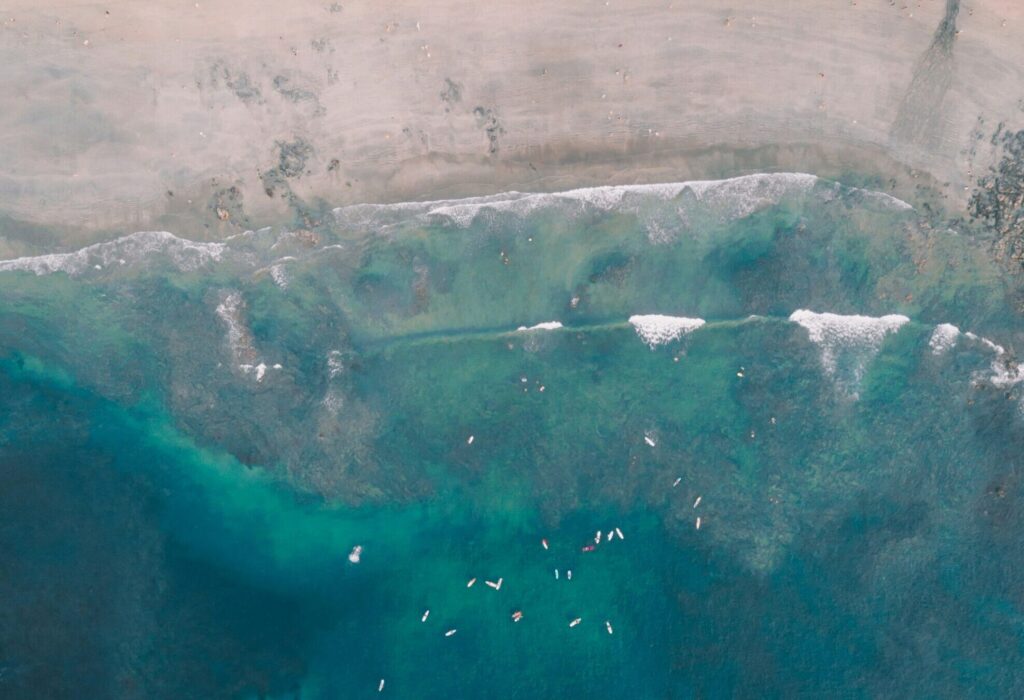 A bird's-eye view of Tamarindo Surf Beach, Costa Rica, showing surfers sitting in ocean, a sandy beach lined with palm trees, and vibrant blue ocean waters