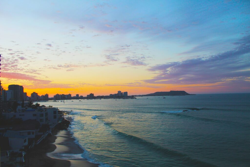 A scenic view of Salinas surf Beach, Ecuador, at sunset, with an apartment building silhouetted against vibrant blue, purple, and yellow skies