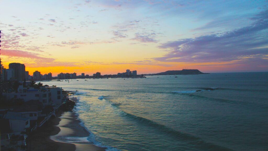 Salinas Beach, Ecuador surf, at sunset with an apartment building silhouetted against blue, purple, and yellow skies and small rolling waves