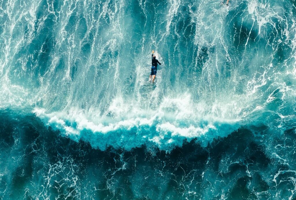 A bird's-eye view of two surfers paddling in blue water, with a wave breaking behind them, bubbles in the ocean