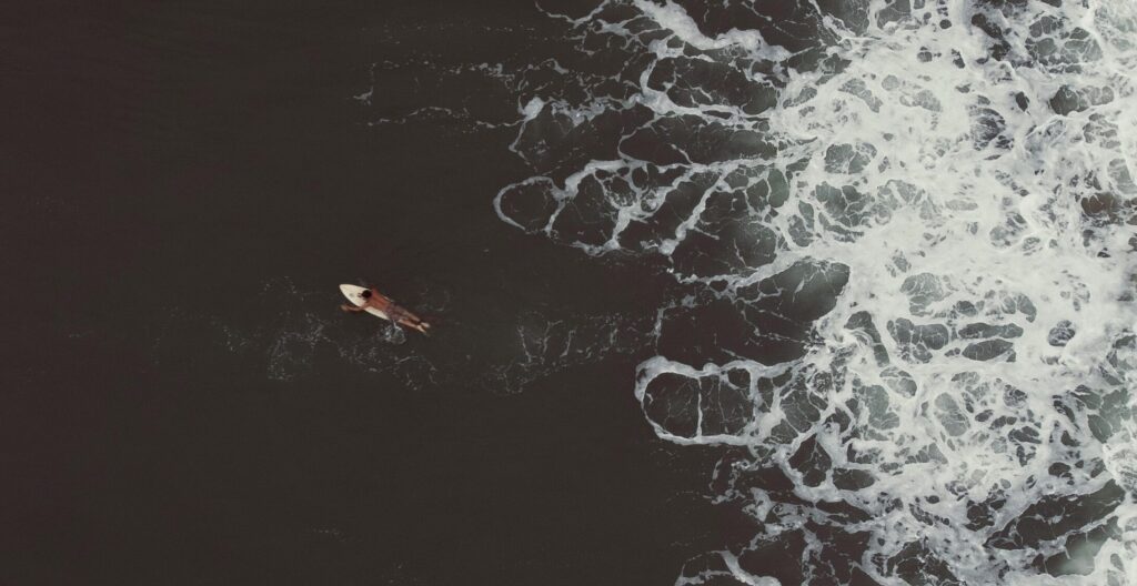 A bird's-eye view of a single man paddling on a surfboard through dark water in Jaco Surf