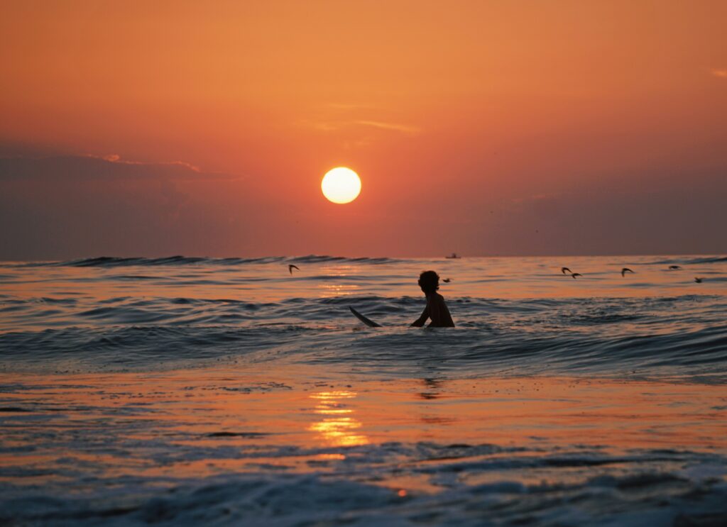 A surfer in arugam bay surf sitting and waiting for a wave, silhouetted against a large sun and vibrant orange skies