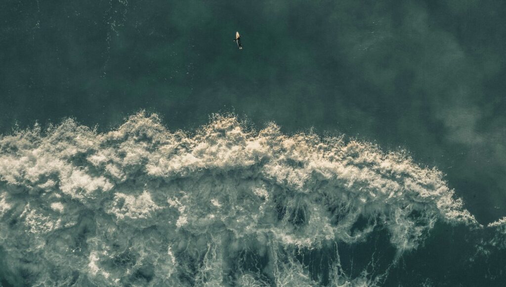 A bird's-eye view of three surfers in a dark green-grey ocean, with a wave breaking behind them