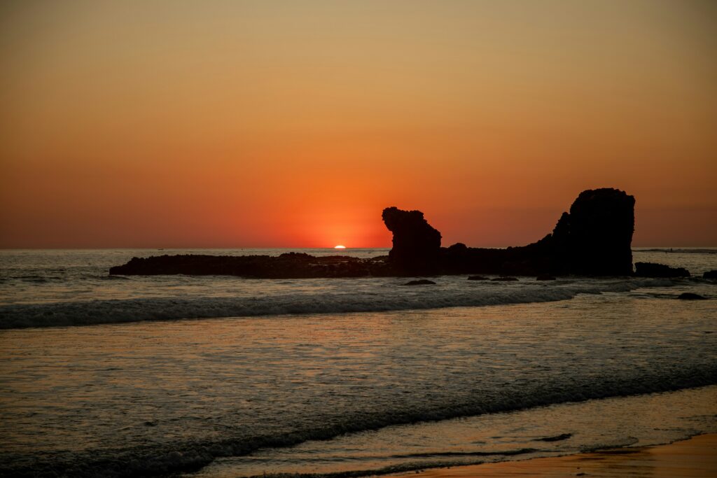 A vibrant sunset at El Tunco surf, El Salvador, with orange and yellow skies over the ocean, showcasing a prominent rock formation in the foreground