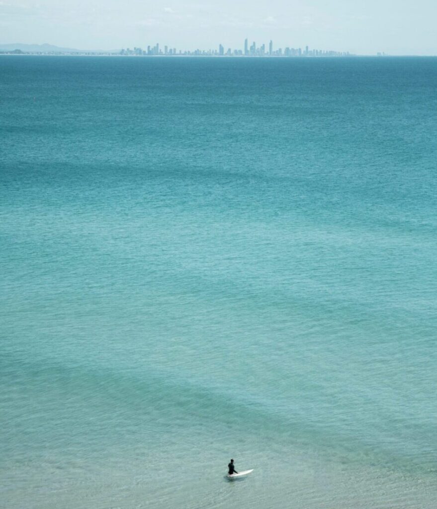 Surfer standing in shallow water with clear blue waves, Gold Coast city skyline in the background under a bright blue sky