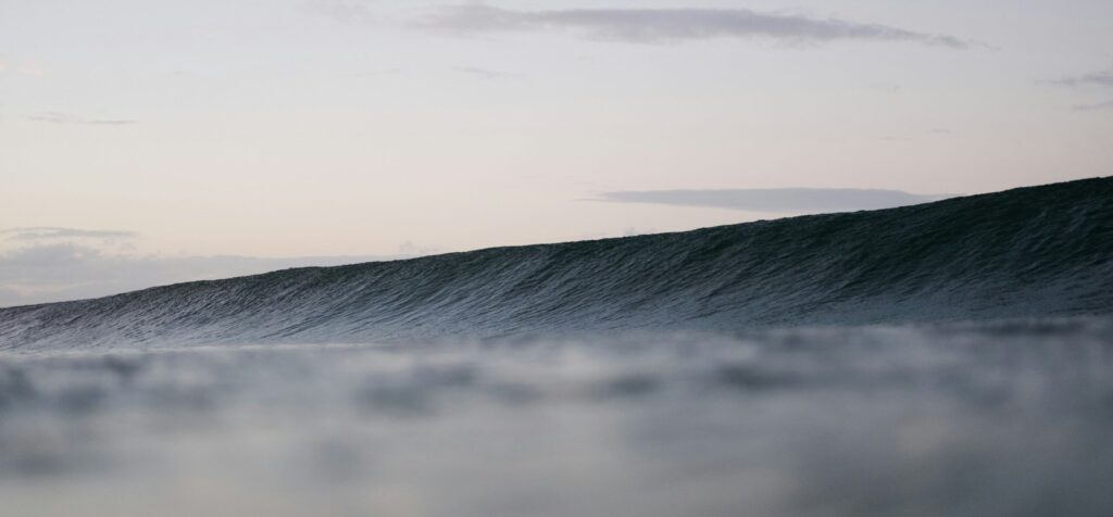 A rolling wave beneath a light blue-grey sky, contrasting beautifully with the deep blue water, creating a serene ocean scene