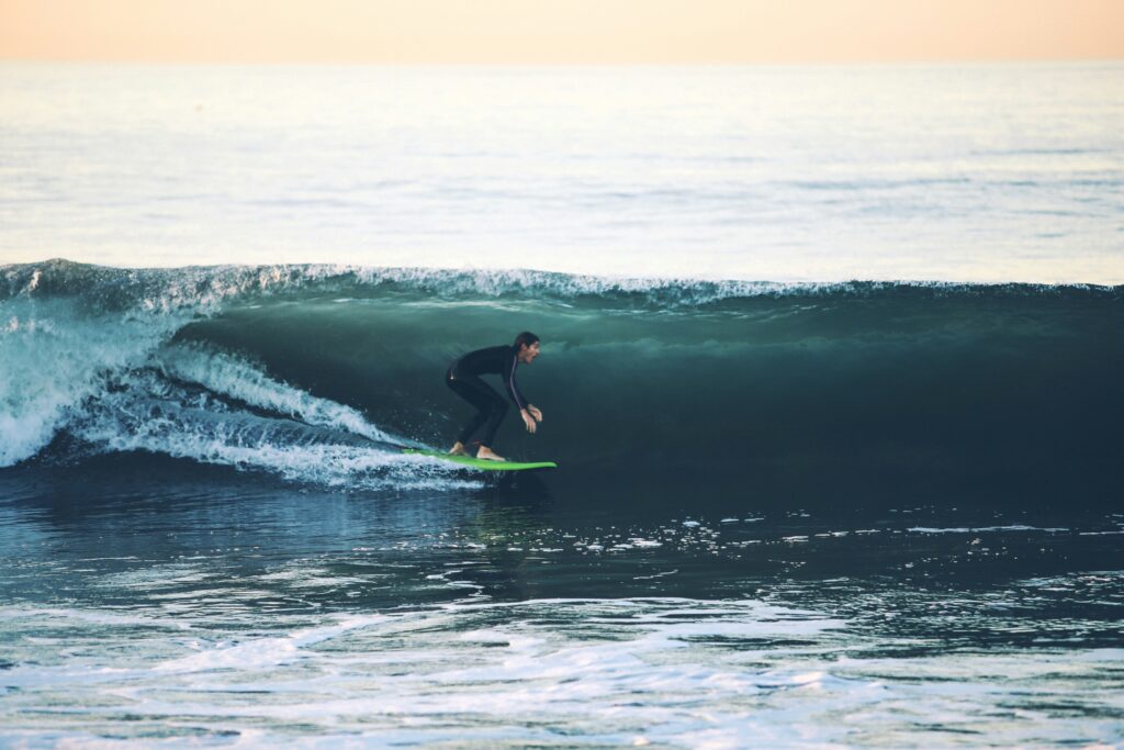 A surfer on a green surfboard rides inside a small left-hand barrel, perfectly tucked as the wave curls, with orange skies glowing in the background