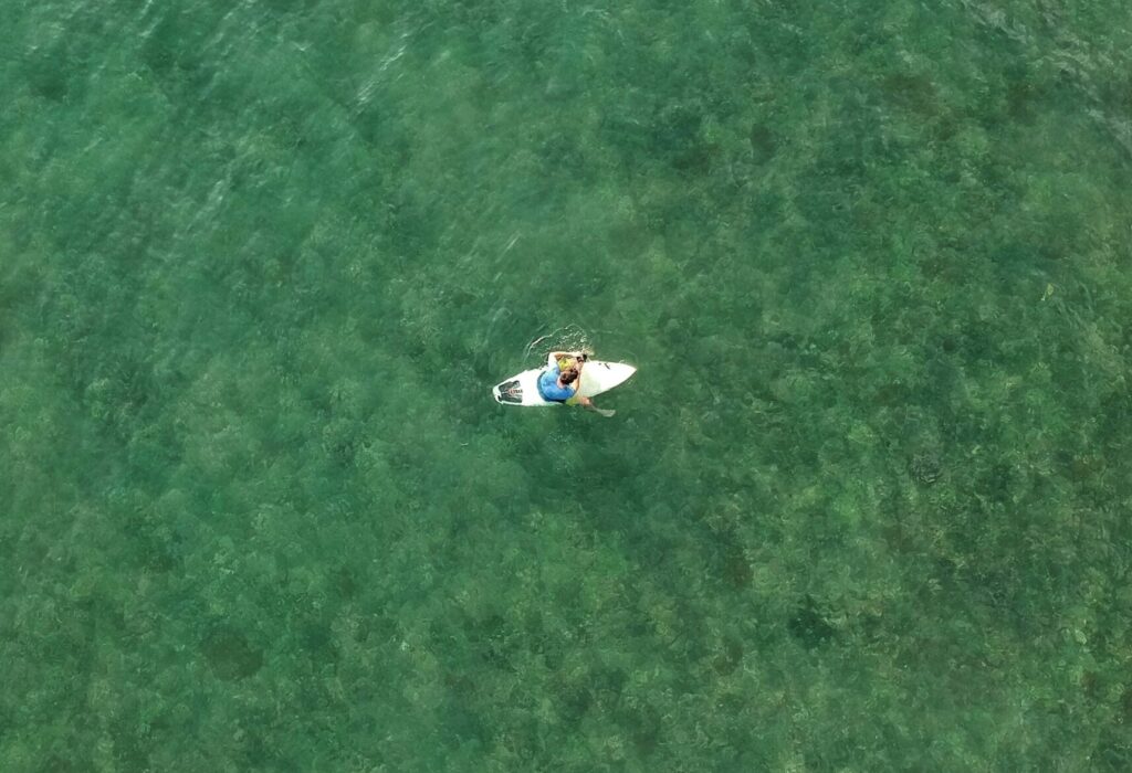 A bird's-eye view of a man sitting alone on his surfboard in clear green-blue water in Puerto Viejo Surf, Costa Rica