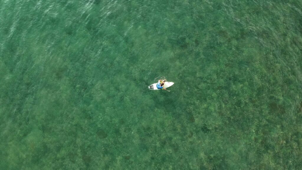 A bird's-eye view of a man sitting alone on his surfboard in clear green-blue water in Puerto Viejo Surf, Costa Rica