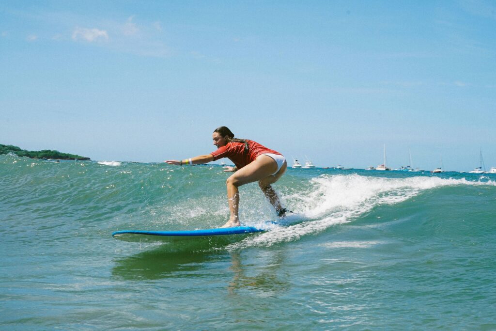 A girl learning to surf on a longboard in Tamarindo, Costa Rica, gliding on gentle waves in warm, clear blue water under bright blue skies.