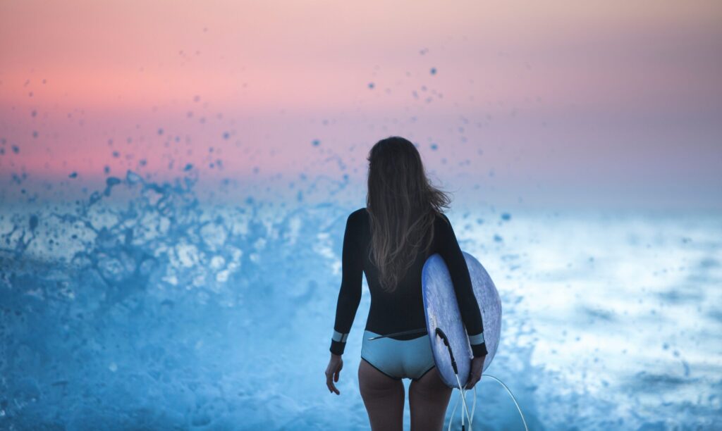 A girl holding a surfboard, poised to enter the water, with waves splashing around her and vibrant orange skies in the background