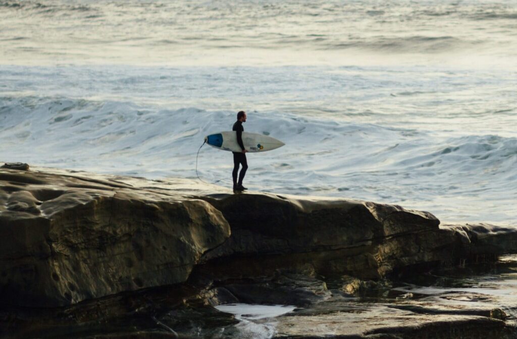 Surfer stands on rocky cliff at Sunset Cliffs surf, San Diego, holding a surfboard under his arm, watching waves crash below under a soft, cloudy sky