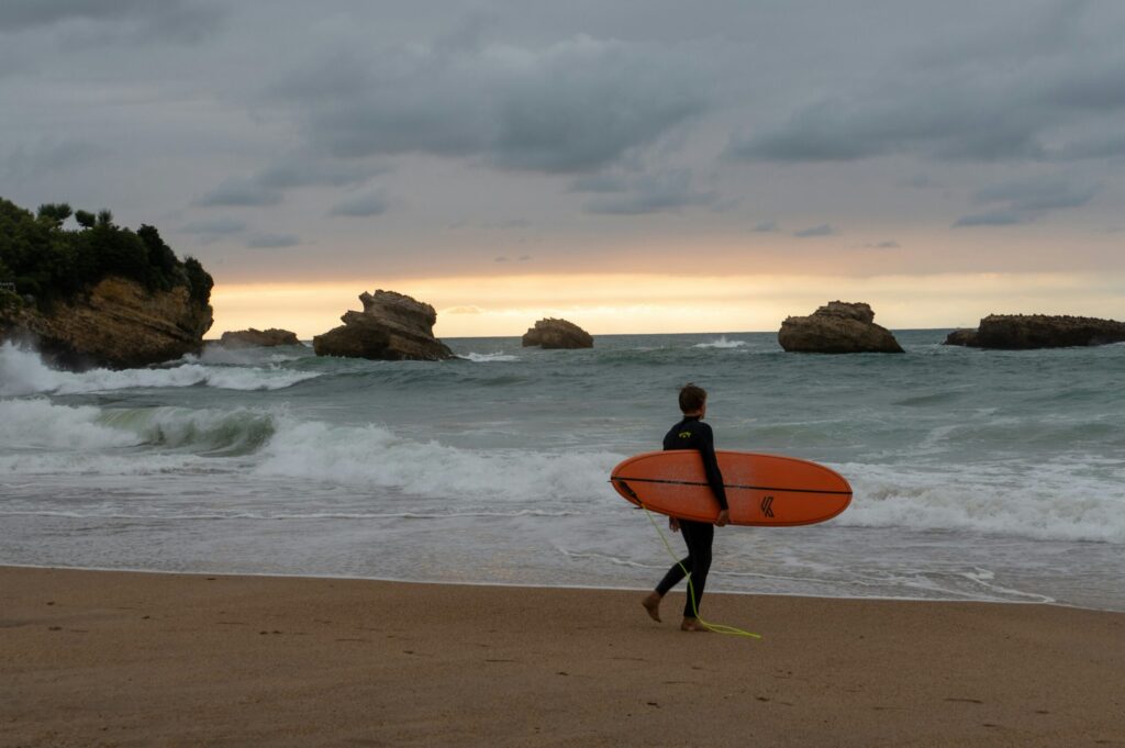 A surfer walking into the ocean in Biarritz surf with their surfboard, preparing for a session, with rocky formations in the background