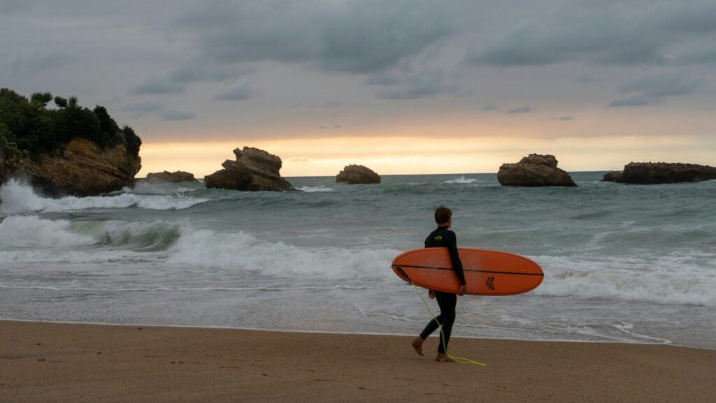 A surfer walking into the ocean in Biarritz surf with their surfboard, preparing for a session, with rocky formations in the background
