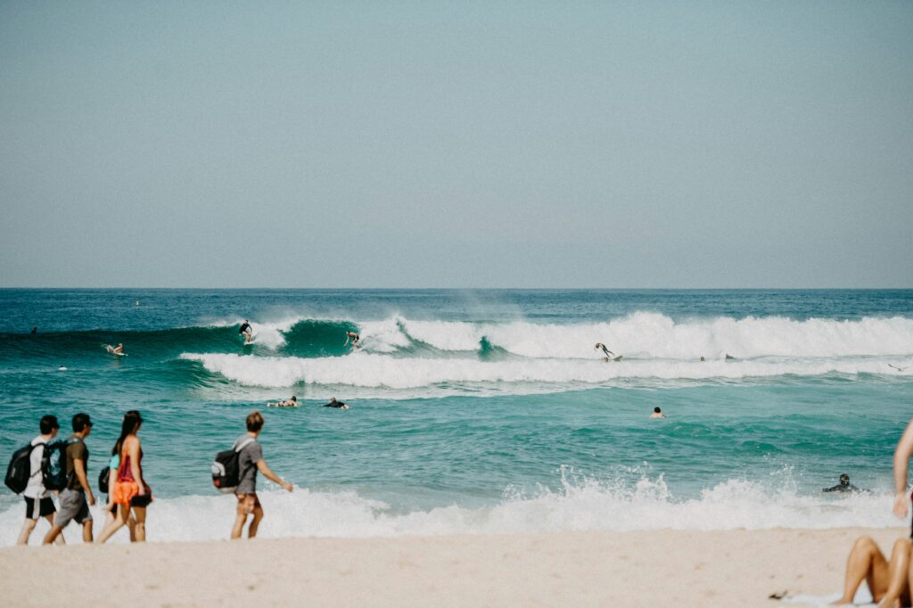 Surfers riding a wave at Bondi Beach, Sydney, with onlookers strolling along the sandy shore