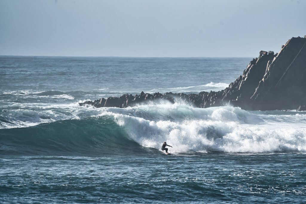 A surfer executes a bottom turn on a left-hand wave in Praia da Arrifana Surf, Algarve, with rocks and clear skies in the background