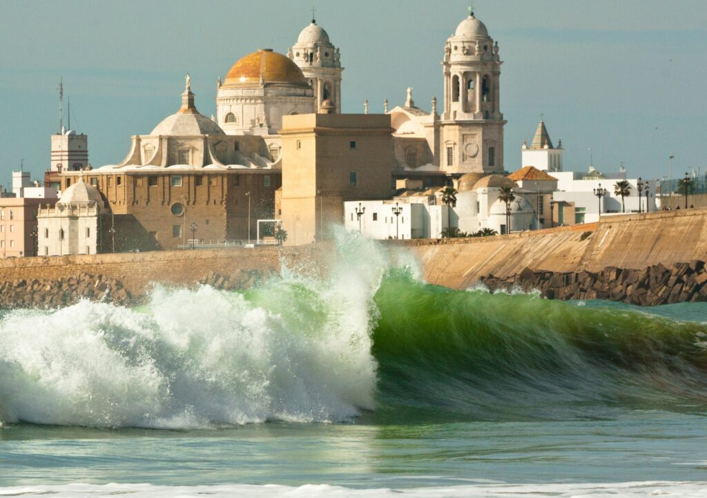 An empty left-hand wave rolls gracefully with Cadiz Surf Cathedral visible in the distant background, under a clear sky