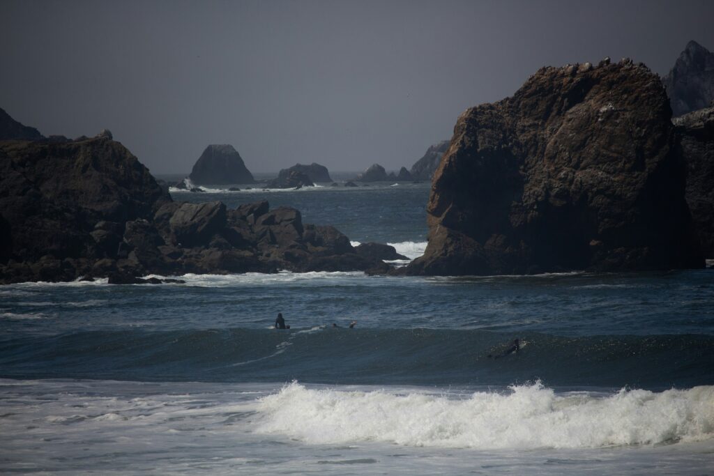 Two surfers are in the water at Pacifica surf, California, with one paddling over a wave. Large rocks emerge from the ocean in the background