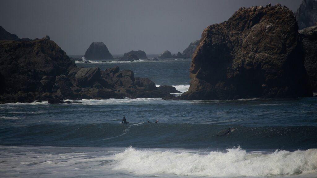 Two surfers are in the water at Pacifica surf, California, with one paddling over a wave. Large rocks emerge from the ocean in the background