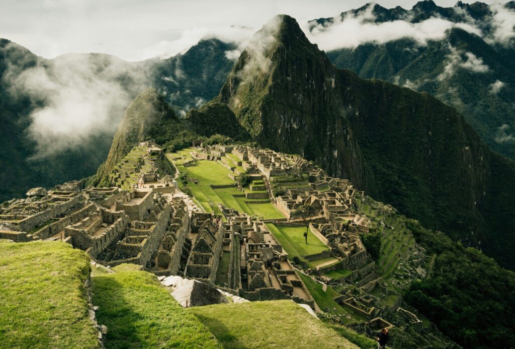 Machu Picchu in Peru, surrounded by green grass and rolling hills, with low clouds drifting over the ancient site