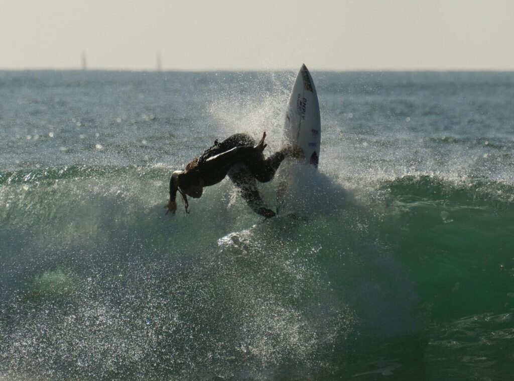 A surfer performs a backside turn on a wave, surrounded by clear skies and vibrant blue-green water