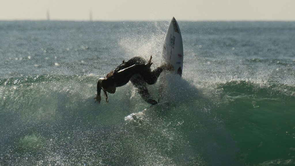 A surfer performs a backside turn on a wave, surrounded by clear skies and vibrant blue-green water