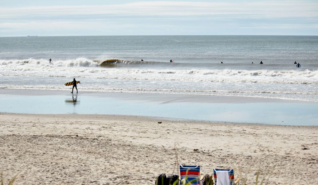 A small barrelling wave seen from the shore as a surfer with a malibu exits the water; two empty beach chairs rest on the sand at Rockaway Beach surf, Queens, NY